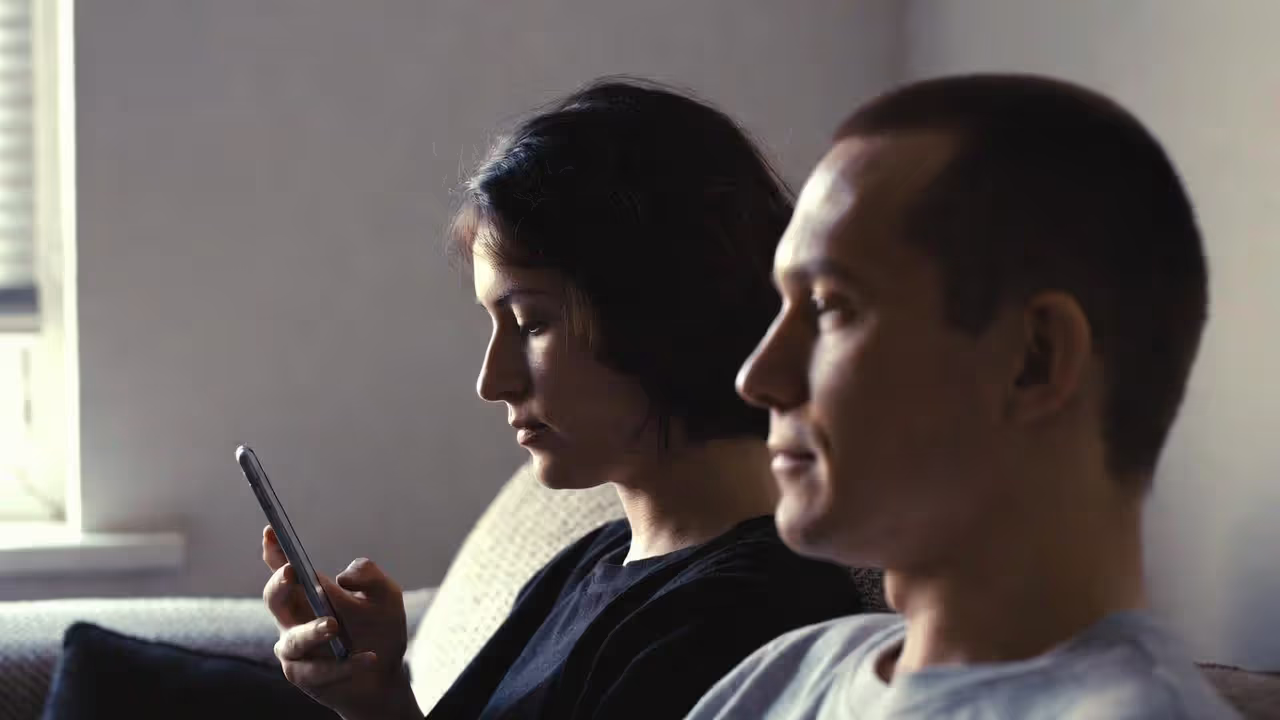 Couple sitting together while one partner looks at phone and the other looks away